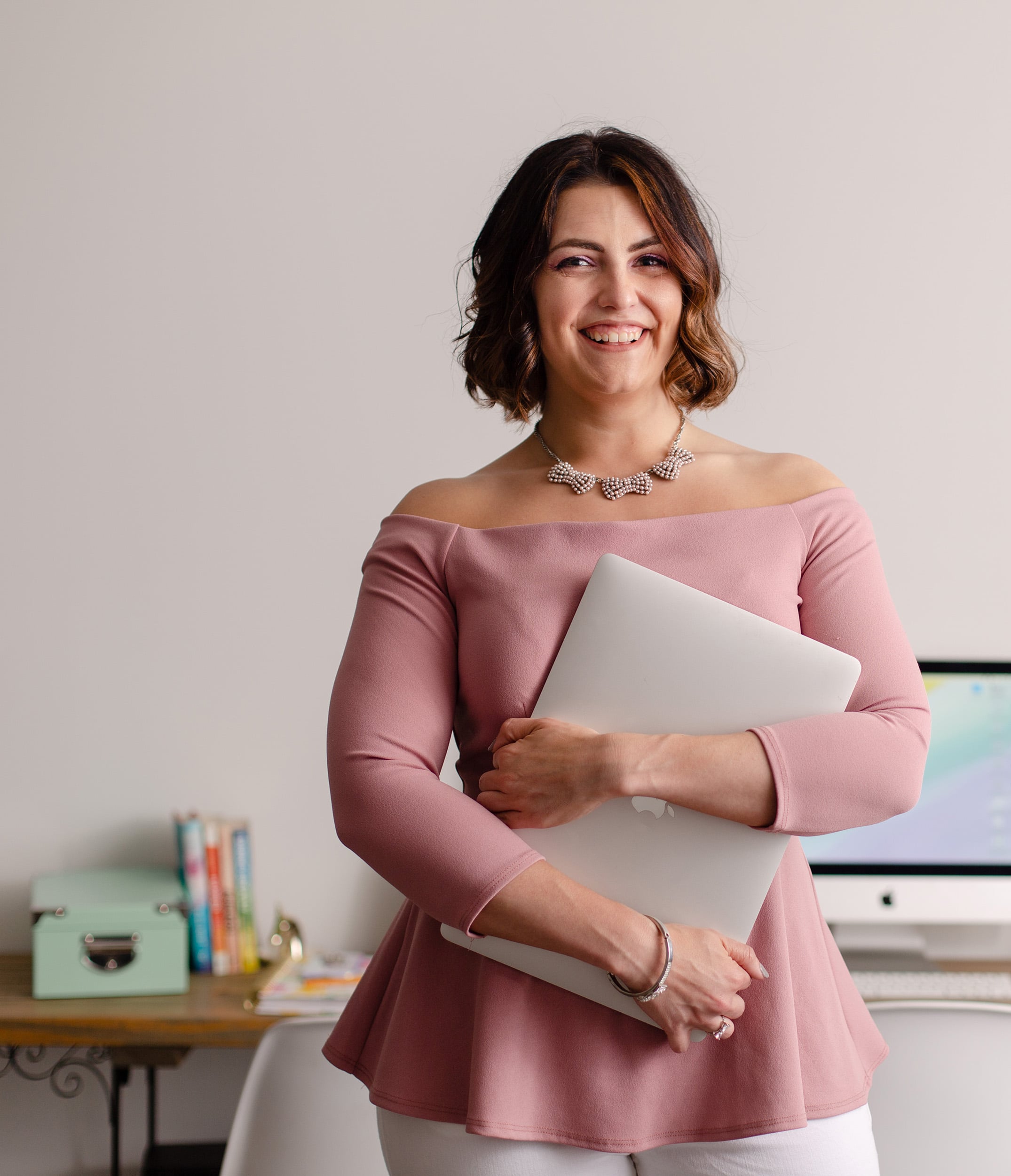 Megan standing as she holds a closed MacBook in front of her, smiling at the camera