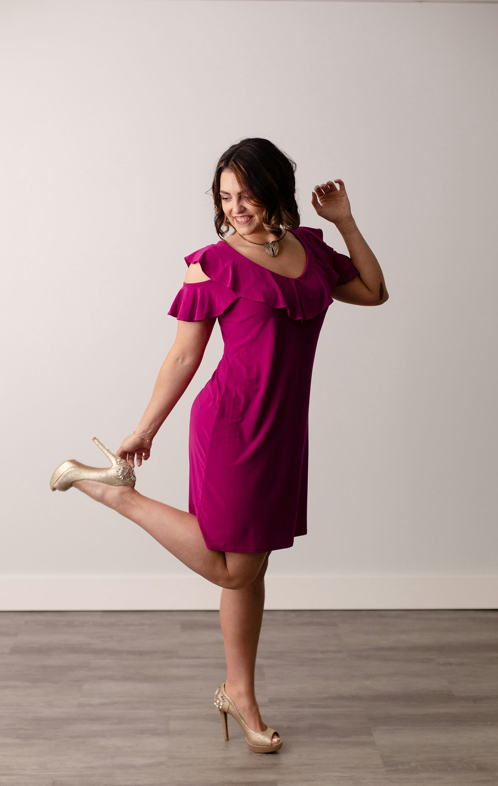 Megan in a loose-fitting purple dress, laughing and looking down while kicking up one foot behind her and touching a gold high heel