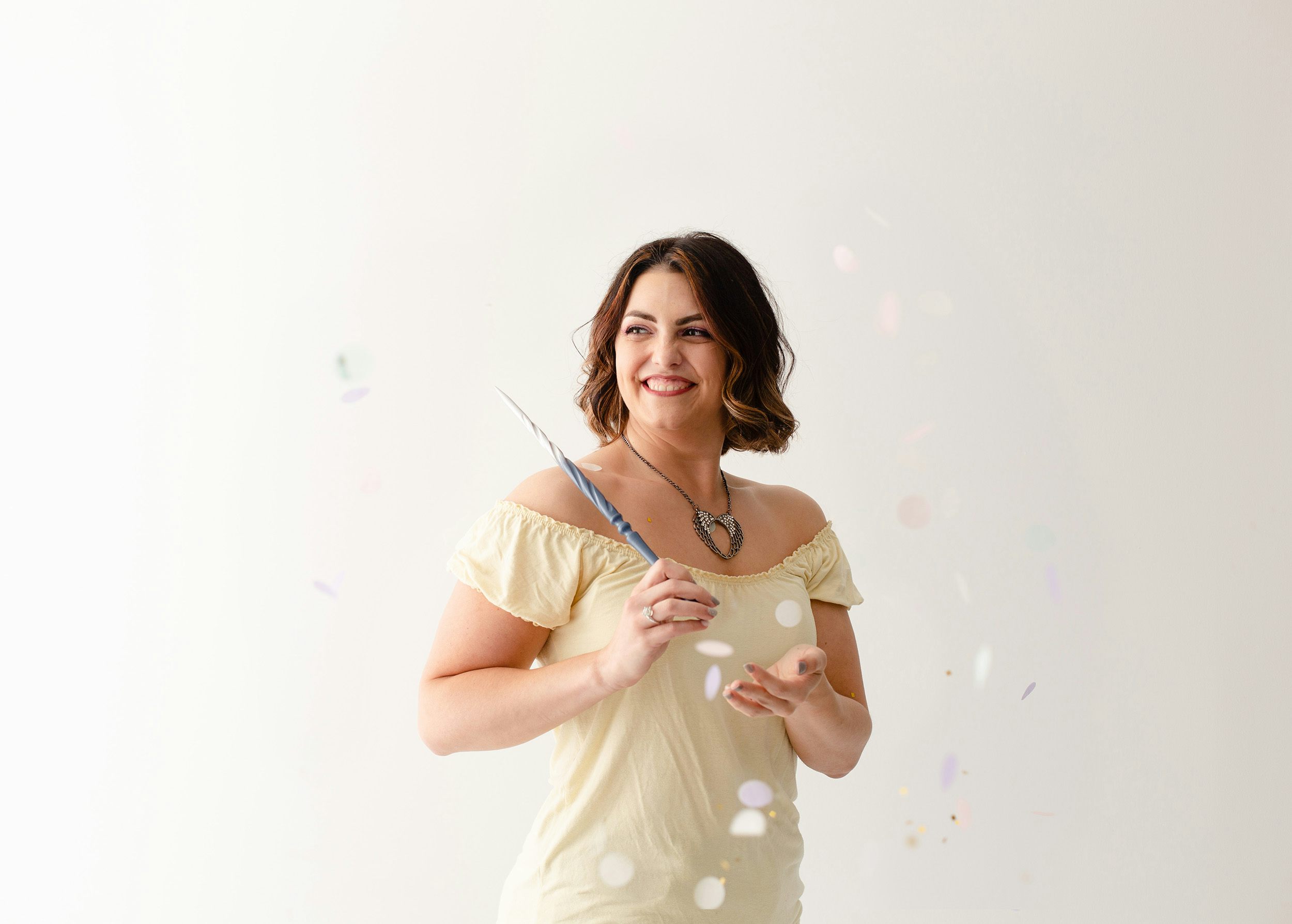 Megan smiling widely as she points a magic wand over her left shoulder, confetti fluttering around her