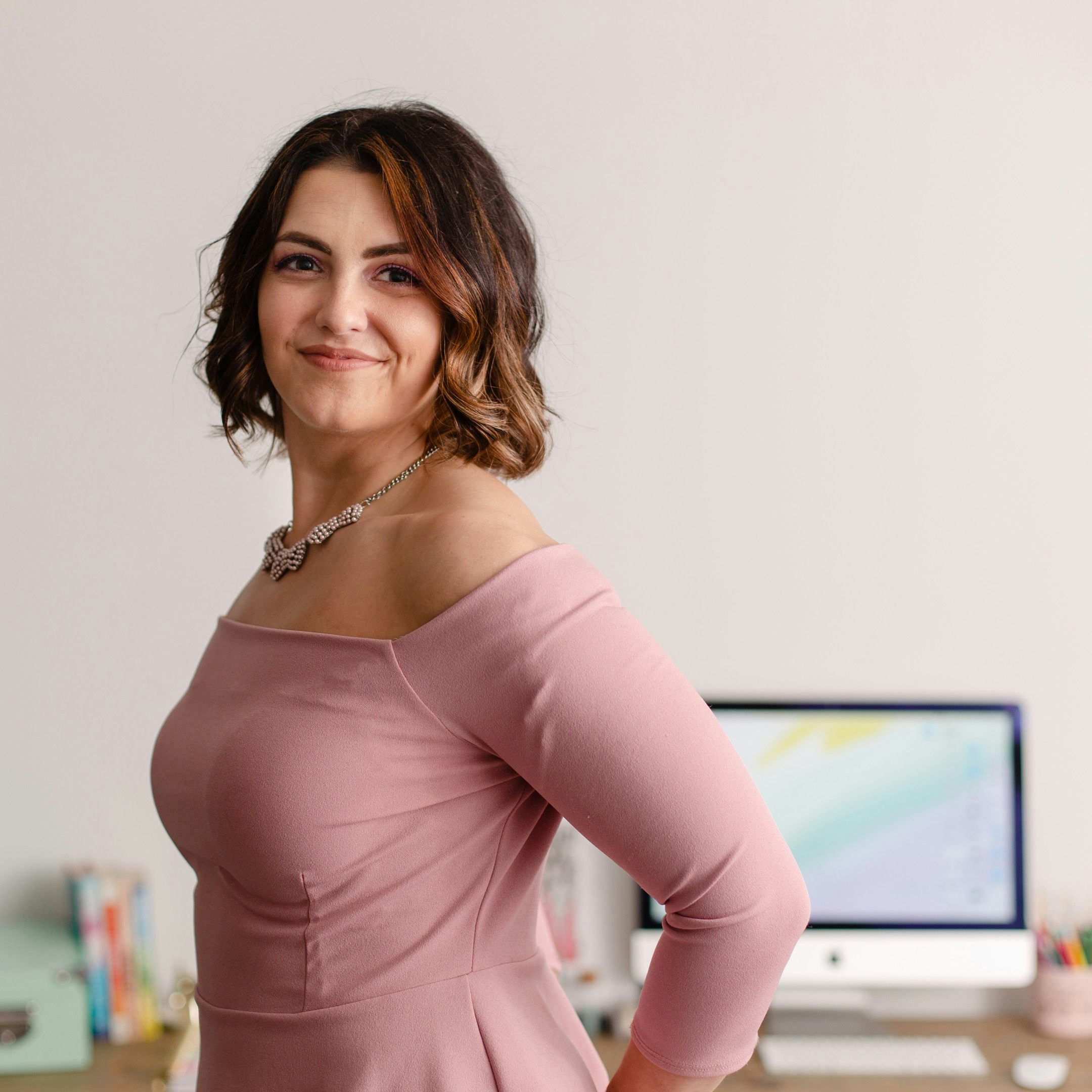 Softly smiling white woman with medium-length dark brown curly hair in a pink blouse, standing in front of a desk with an iMac